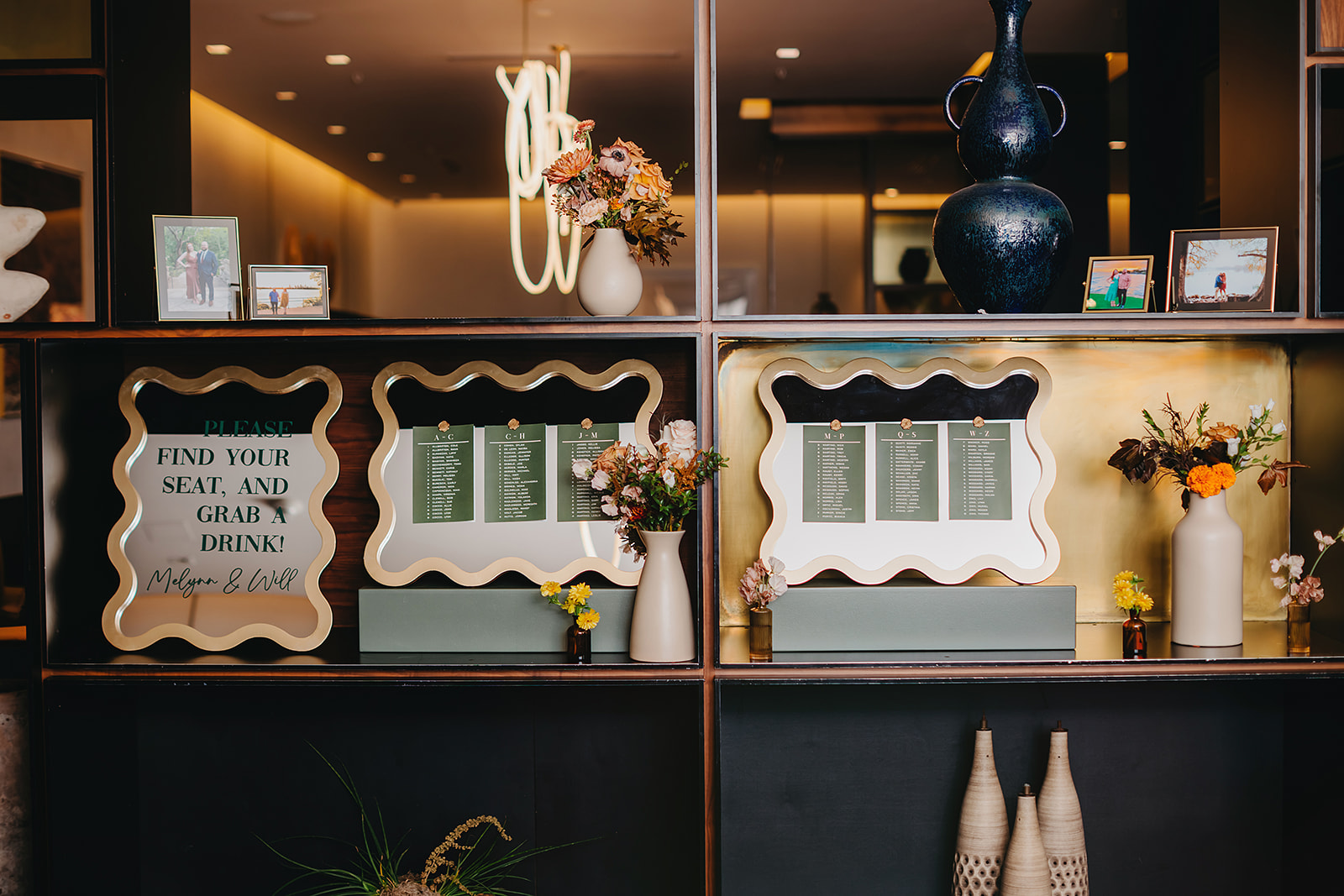 Shelves display framed seating charts, flowers, vases, and small photo frames in a warmly lit interior space with decorative accents.