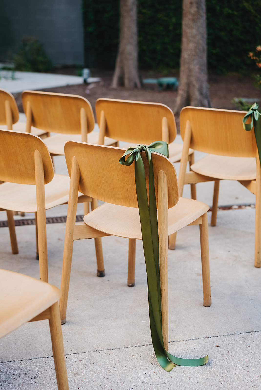 Rows of light wooden chairs are arranged outdoors, each decorated with a long, green ribbon tied in a bow on the backrest.