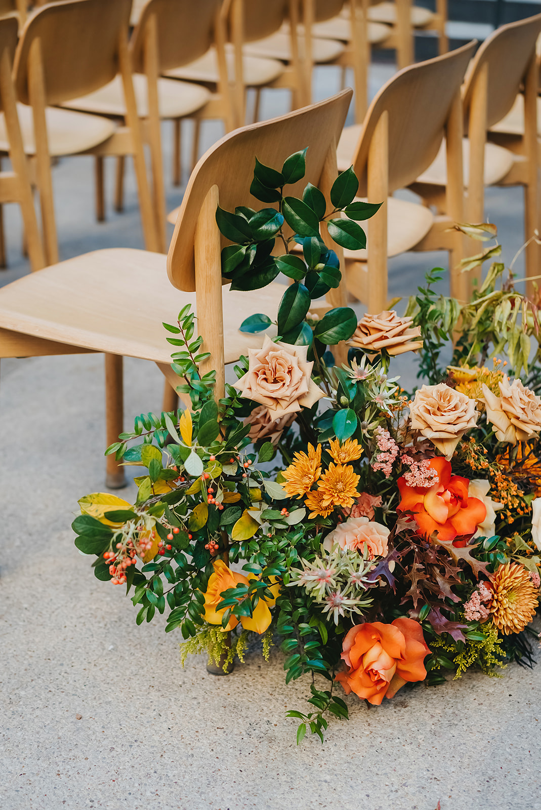 Wooden chairs are arranged in rows on a concrete floor, with a large, colorful floral arrangement featuring orange and peach flowers and green foliage beside one chair.