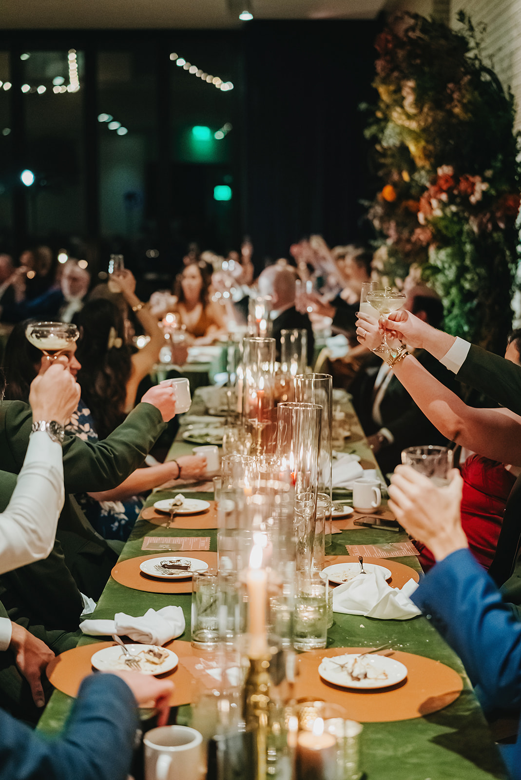 A group of people sit around a long, decorated reception table raising glasses in a toast with candles and plates of dessert, the perfect representation of the elevated guest experience In Ink Weddings creates through design.