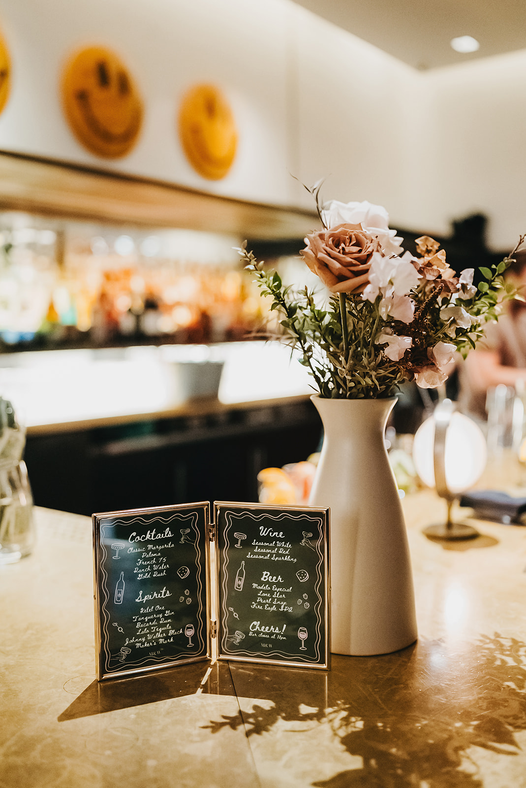 A flower vase and a framed cocktail and wine menu sit on a bar counter with a blurred background of bottles and warm lighting.