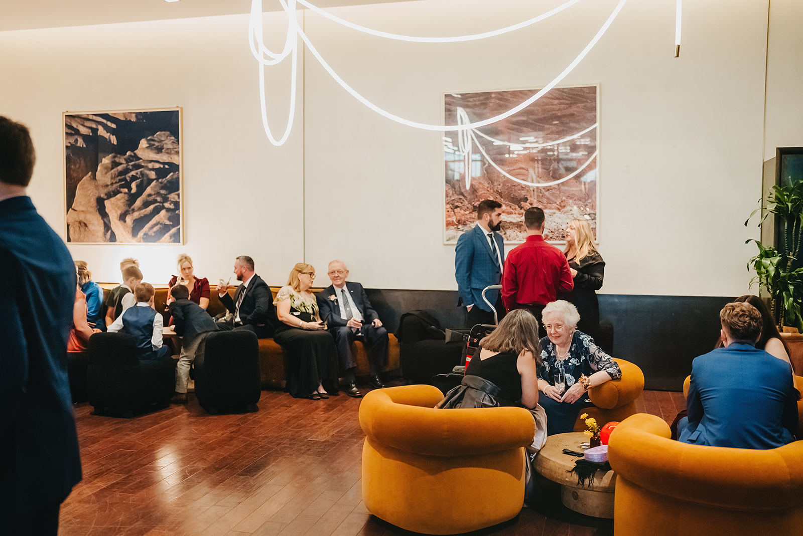 People sit and socialize in a modern lounge area with orange chairs, art on the walls, and bright overhead lighting, showing how In Ink Weddings creates an elevated guest experience through design and details.