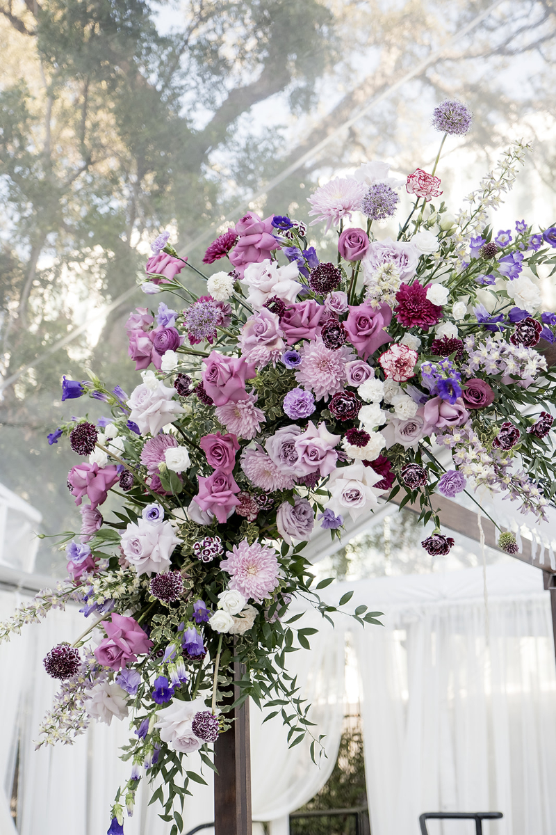 An arrangement of purple, lavender, white, and burgundy flowers decorates a wooden structure, with outdoor trees and sheer white drapery in the background.