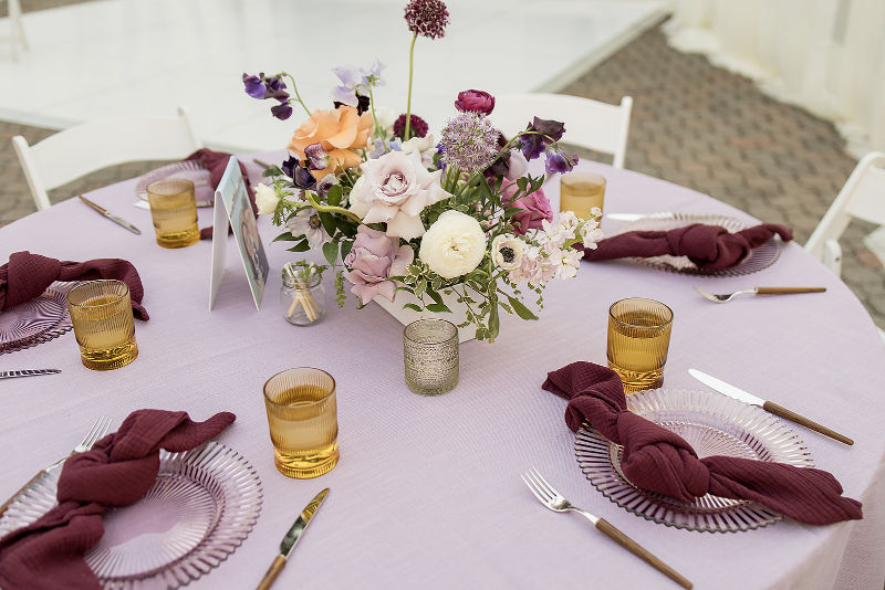 A round table with a lavender tablecloth, purple floral centerpiece, amber glasses, clear plates, silverware, and burgundy napkins set for six people.