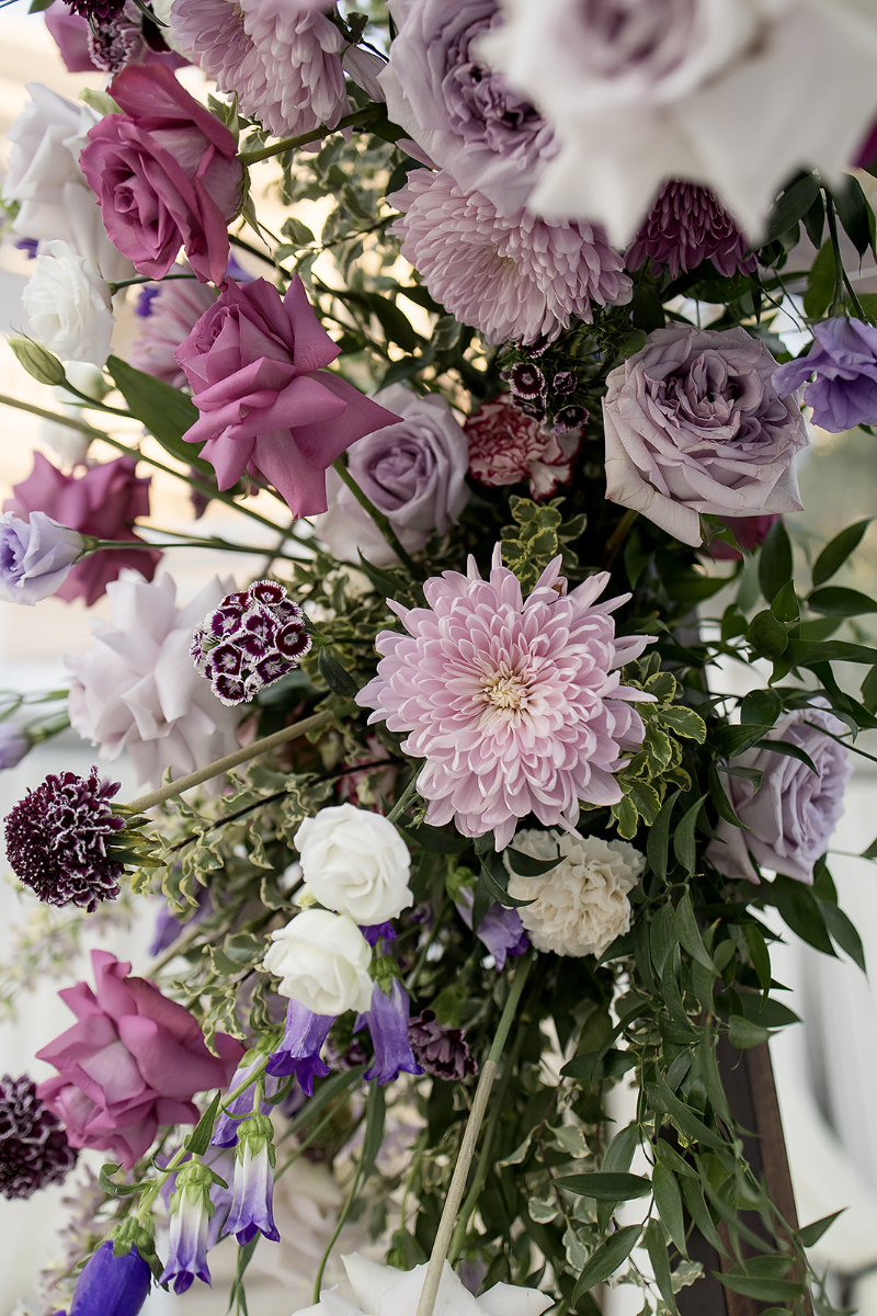 A close-up of a floral arrangement featuring pink and purple roses, chrysanthemums, white lisianthus, and green foliage.