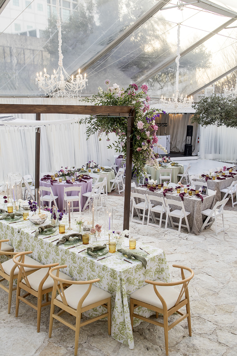 The Allan House Austin decorated for wedding with elegant chandeliers, draped ceiling, floral centerpieces, and tables set with patterned linens, glassware, and wooden chairs under a clear tent.