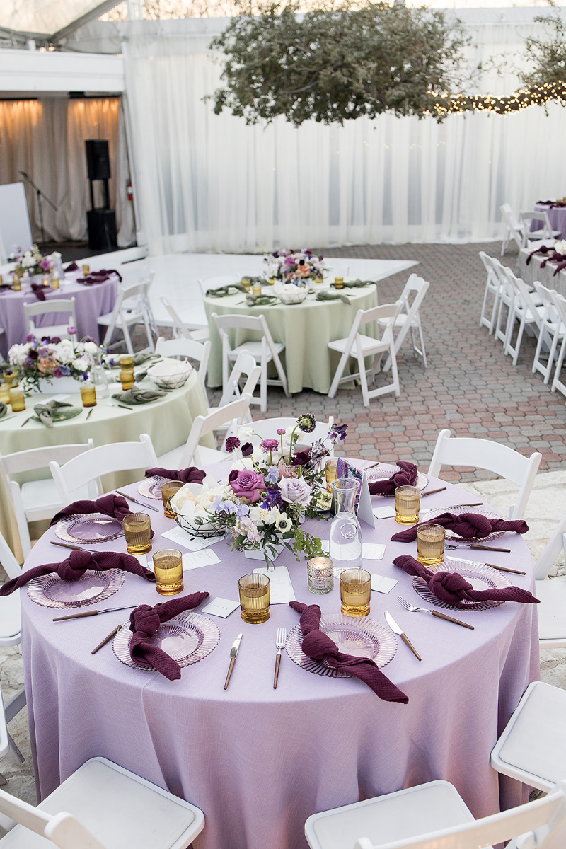 Elegant outdoor event setup with round tables covered in pastel purple and green tablecloths, floral centerpieces, and neatly arranged plates, glasses, and utensils.