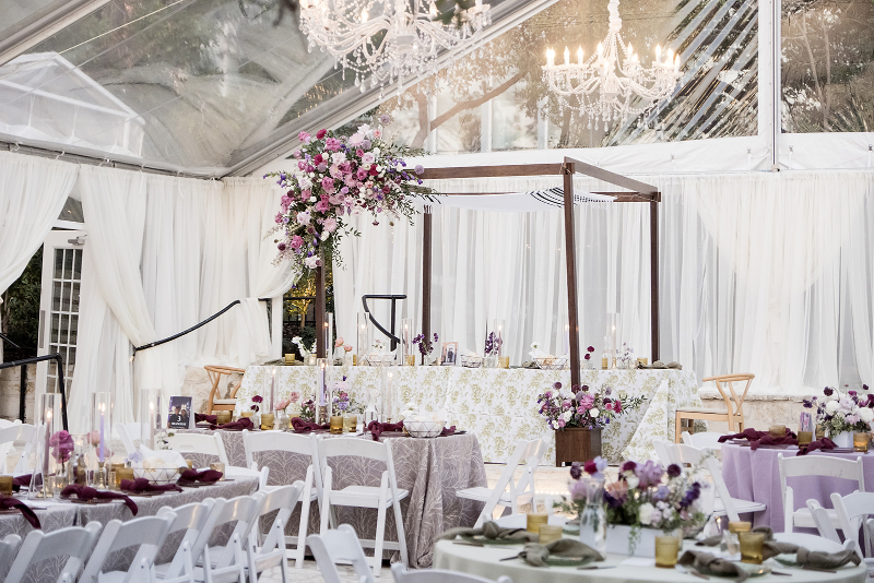 Elegant wedding reception setup under a clear tent with chandeliers, white drapery, floral centerpieces, and decorated tables and chairs.