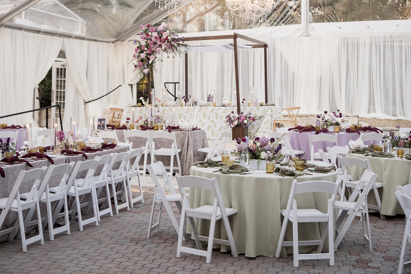A formal event setup with round and rectangular tables covered in pastel linens, floral centerpieces, white chairs, and a flower-adorned canopy under a clear tent.