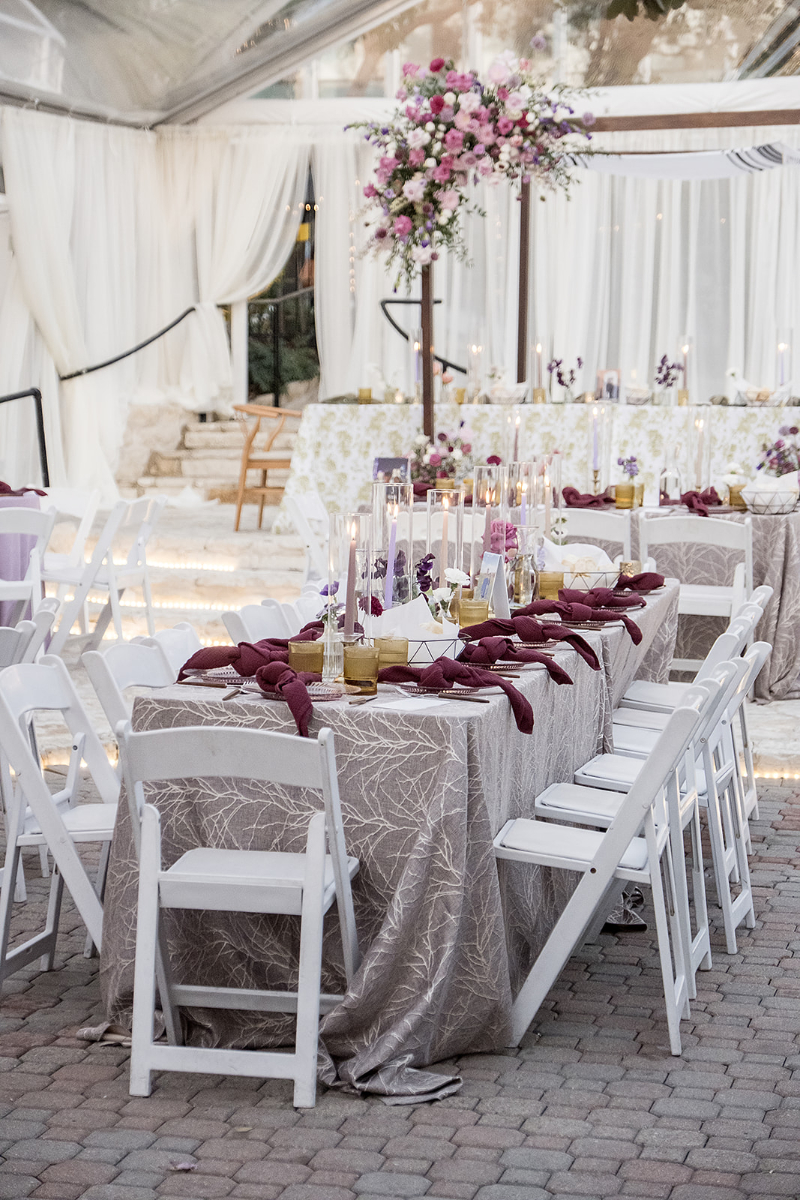 Elegant outdoor event setup with tables covered in patterned cloths, white chairs, maroon napkins, candles, and a tall floral centerpiece with pink and purple flowers.