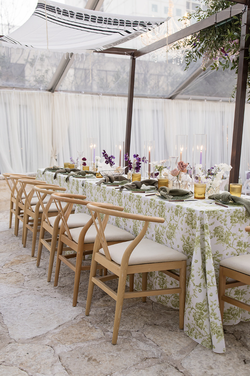 A long dining table with a green and white floral tablecloth, set with candles, floral arrangements, and wooden chairs in an outdoor tented space.