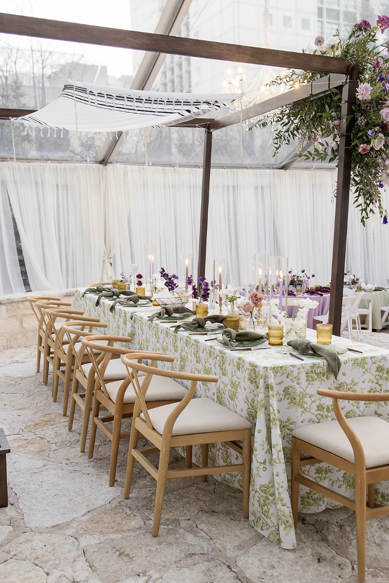 A long dining table with wooden chairs, floral tablecloths, candles, and purple floral centerpieces set under a canopy in a bright, enclosed outdoor space.