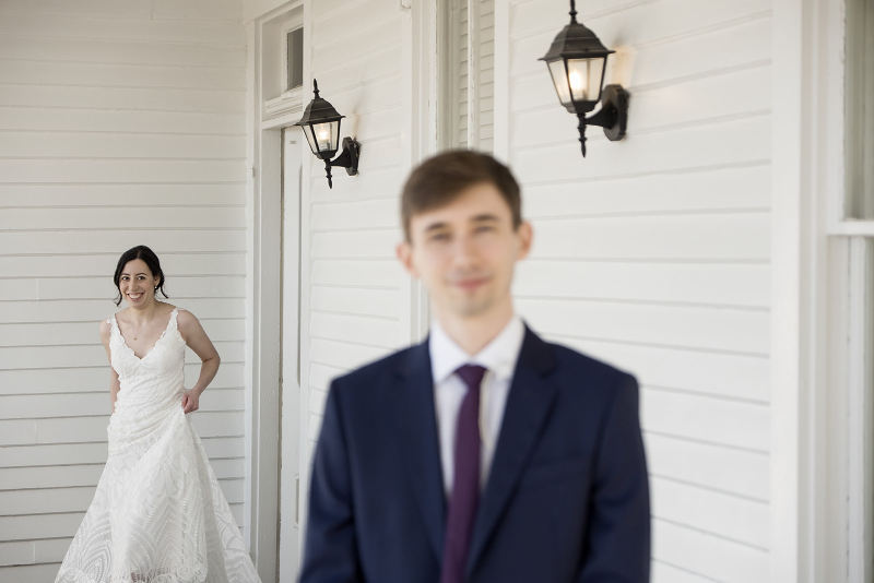 A bride in a white dress stands in the background smiling, while a man in a dark suit stands in the foreground on a white porch with wall-mounted lamps.