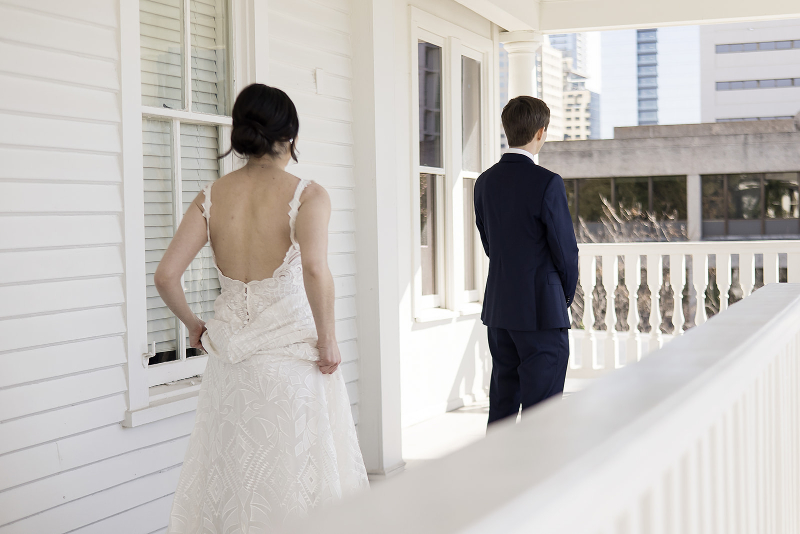 A bride in a white dress stands on a porch facing a groom in a dark suit, who is turned away and looking toward the city buildings in the background.