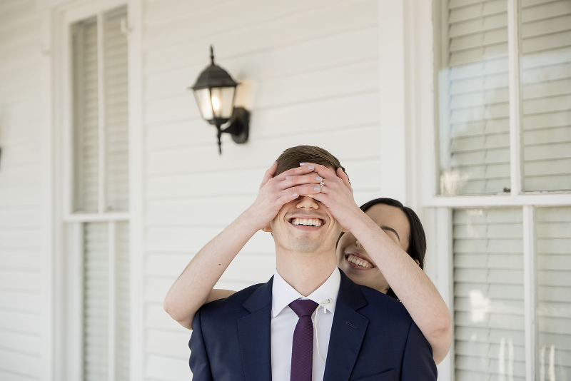 A woman playfully covers a man's eyes from behind as he smiles, both dressed formally, standing in front of a white house with shuttered windows.