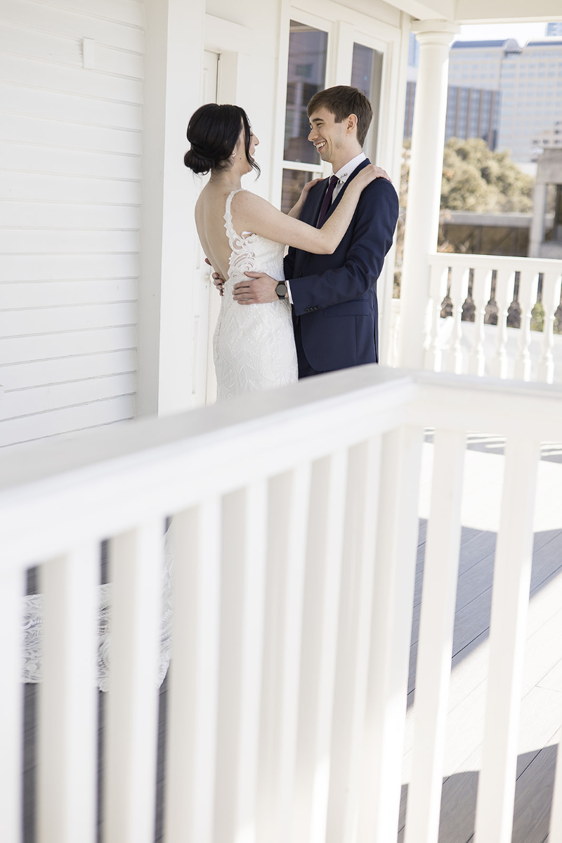 A bride and groom stand on the porch of The Allan House Austin facing each other and smiling, with the bride in a white dress and the groom in a dark suit.