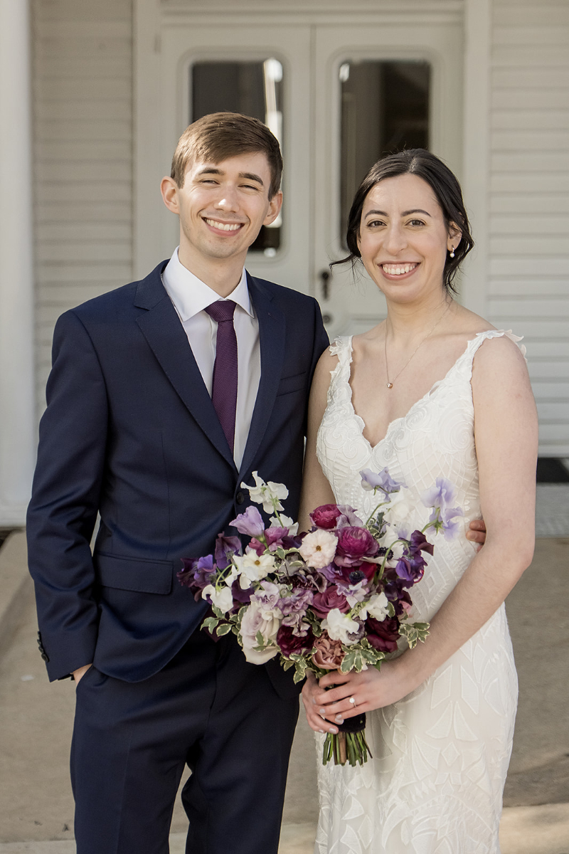 A man in a navy suit and a woman in a white dress holding a bouquet of purple and white flowers stand smiling in front The Allan House Austin.