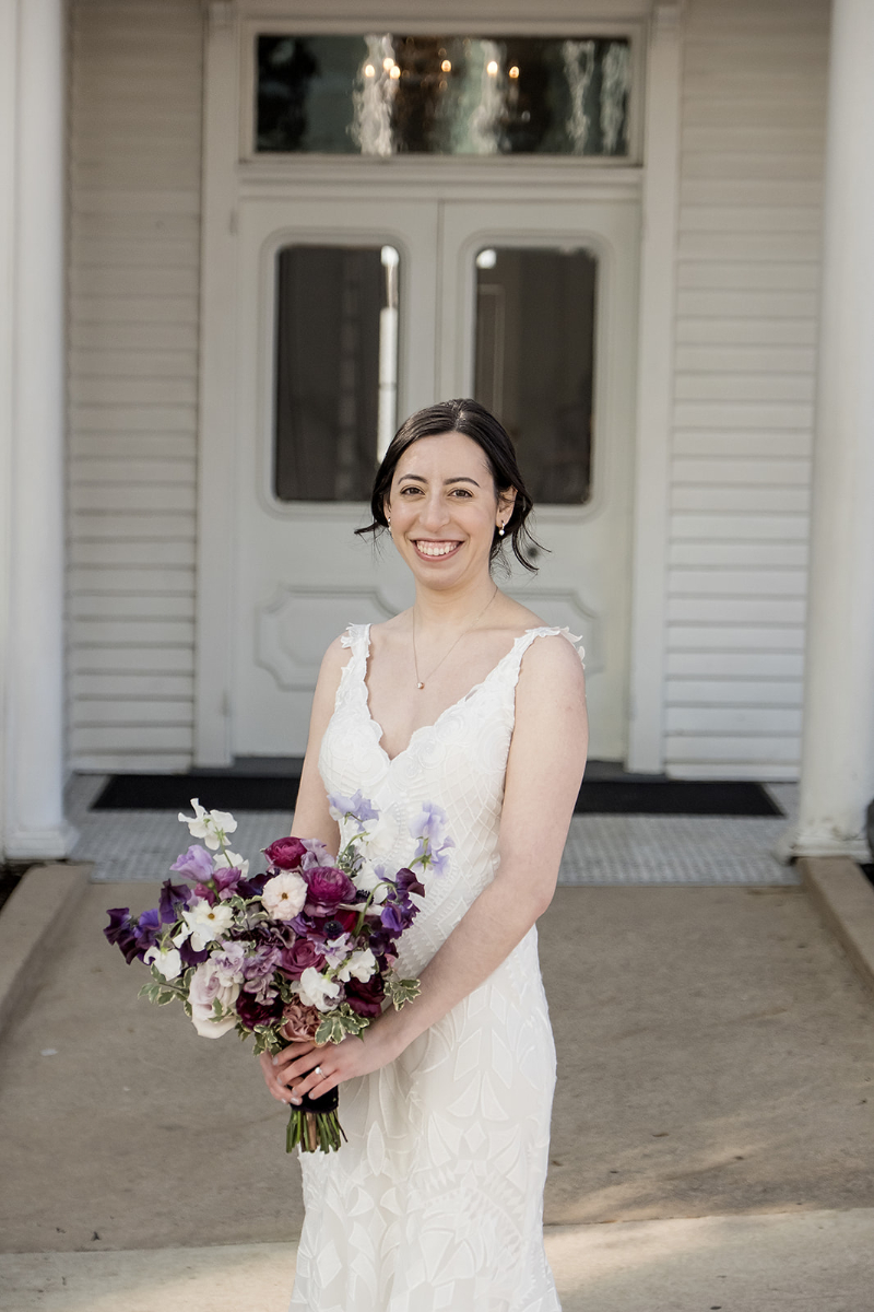 A woman in a white sleeveless wedding dress holds a bouquet of purple and pink flowers while standing in front of a white building with double doors.