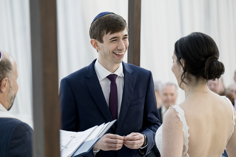 A man in a suit smiles while holding a ring in front of a woman in a white dress during a wedding ceremony, with an officiant holding documents nearby.