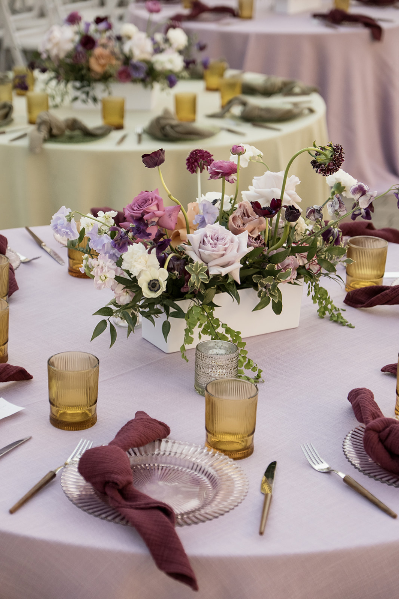 Round table with a lavender tablecloth, purple floral centerpiece, yellow glassware, clear plates, gold utensils, and burgundy napkins; other similarly set tables in the background.