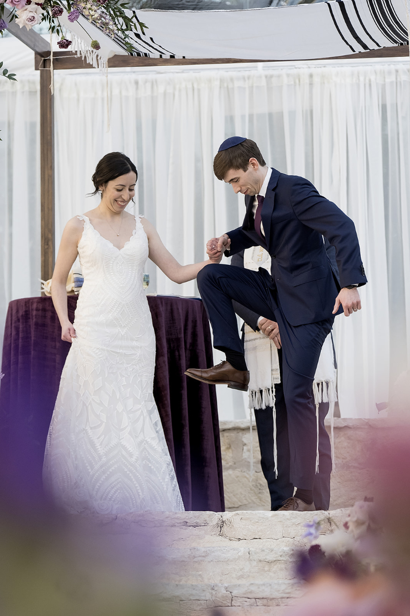 A bride and groom stand under a canopy as the groom prepares to break a glass with his foot during a wedding ceremony.