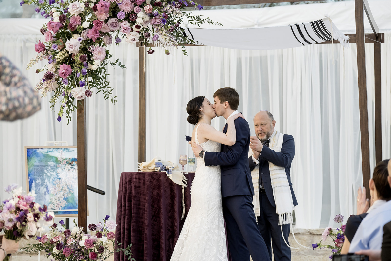 A bride and groom kiss under a floral arch at their wedding ceremony while an officiant stands behind them, clapping.