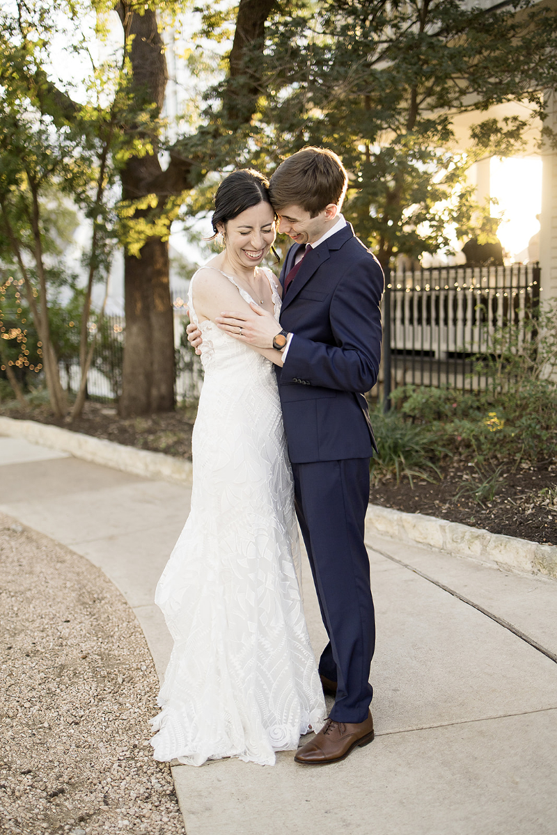 A bride in a white dress and a groom in a navy suit embrace and smile outdoors on a sunlit path, surrounded by trees and greenery.
