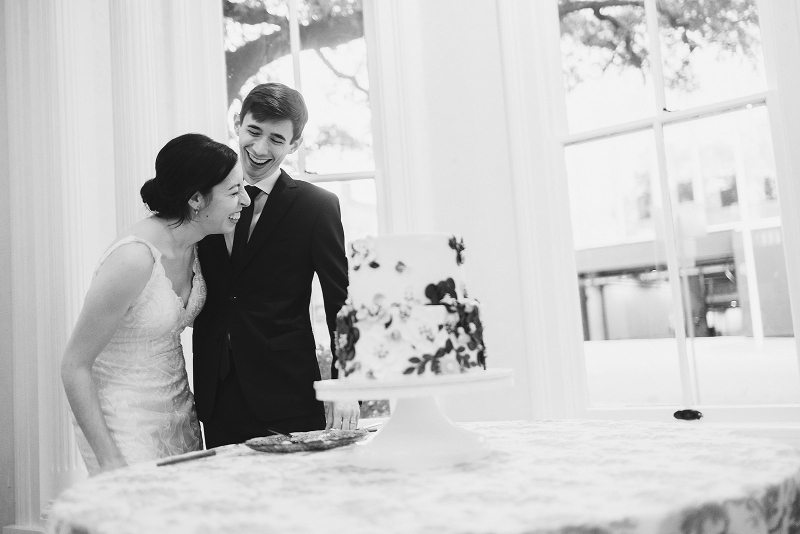 A bride and groom stand by a decorated cake on a table, smiling and leaning toward each other in a bright room with large windows.