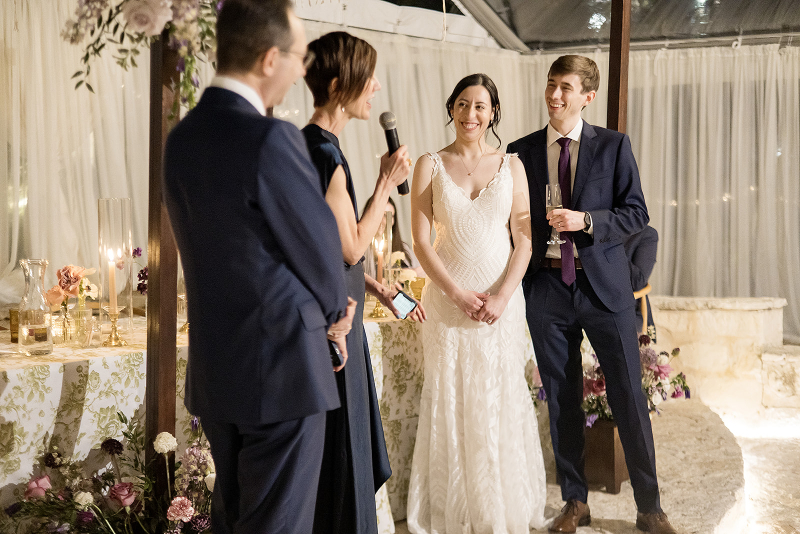 Four people dressed formally stand together at a wedding reception. One woman speaks into a microphone while the bride and groom listen and smile.