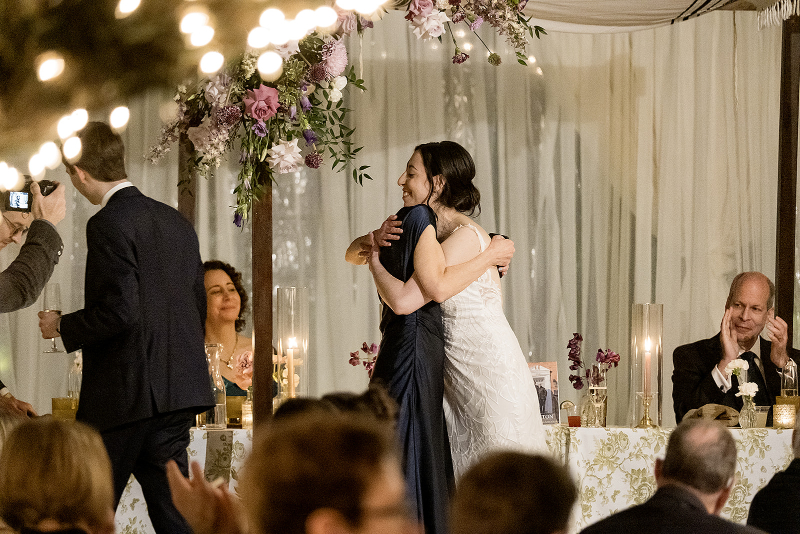 A bride and another woman hug under a floral arch at a wedding reception, while guests sit at a decorated table and others look on.