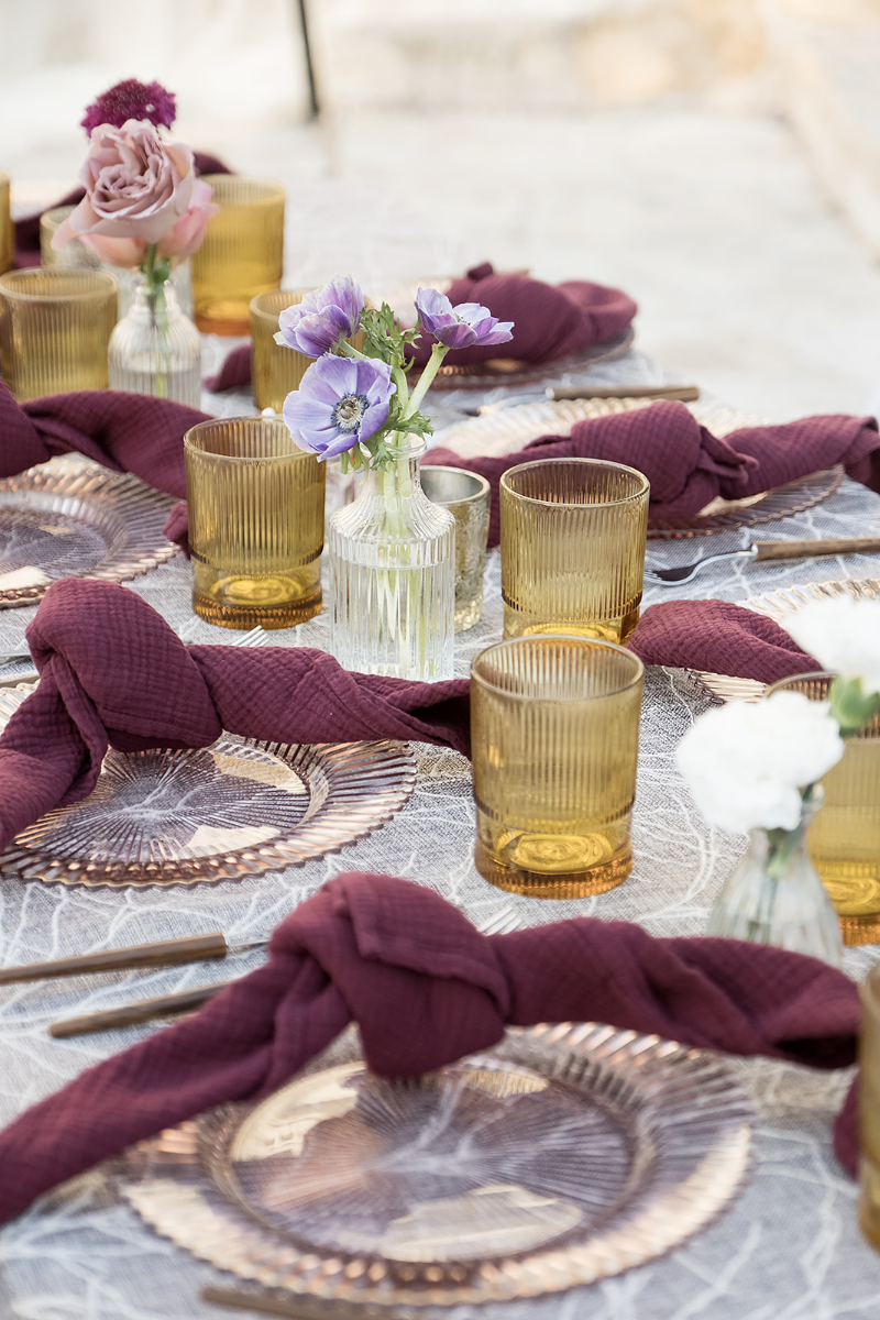 Table setting with clear plates, amber glasses, mauve napkins tied in knots, and small vases holding purple and white flowers on a patterned tablecloth.