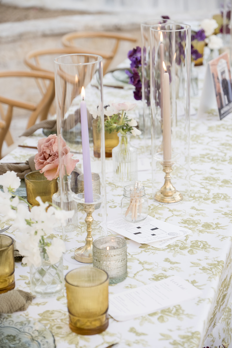 A close-up of a decorated table with floral arrangements, candles in glass holders, glassware, and printed menus on a white and green patterned tablecloth.