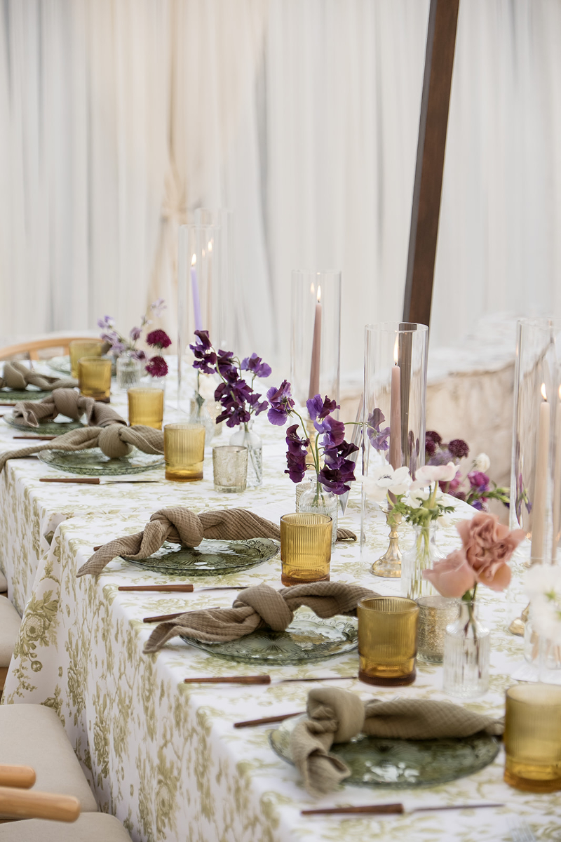 A decorated dining table with floral-patterned tablecloth, purple and pink flowers in vases, yellow glassware, green leaf plates, and beige napkins tied in knots.