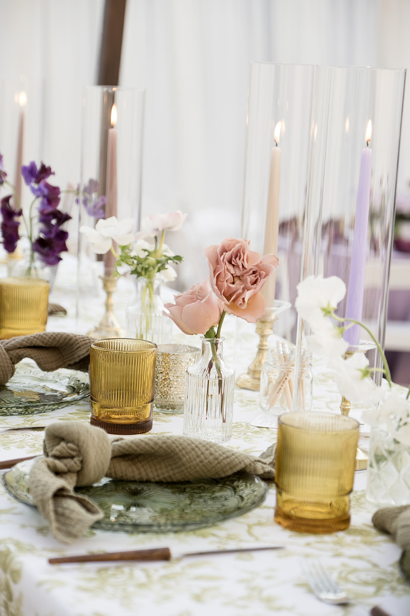 Elegant table setting with yellow glassware, green napkins, floral-patterned plates, pink and white flowers in vases, and lit taper candles in glass holders.