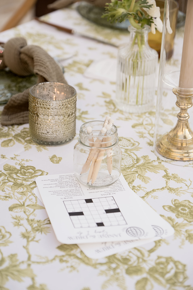 A small glass jar holding pens sits on top of a crossword puzzle sheet on a floral tablecloth, surrounded by a candle, candlestick, and a vase with flowers.
