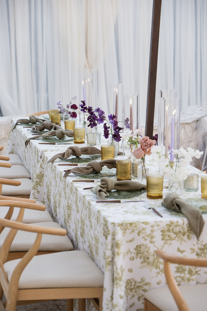 A long dining table with a floral tablecloth is set with yellow glasses, plates, napkins, purple and white flowers, and tall glass candle holders. Wooden chairs are arranged along the sides.