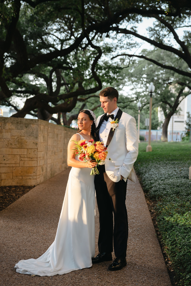 A bride in a white dress holds a bouquet of flowers while standing beside a groom in a white tuxedo jacket on a tree-lined path at sunset.