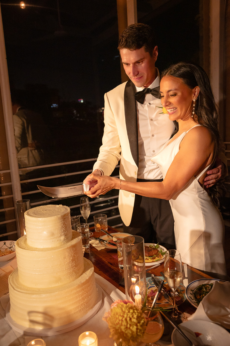 A couple in formal attire stands together, smiling as they cut a three-tiered white cake at their Santa Barbara wedding.