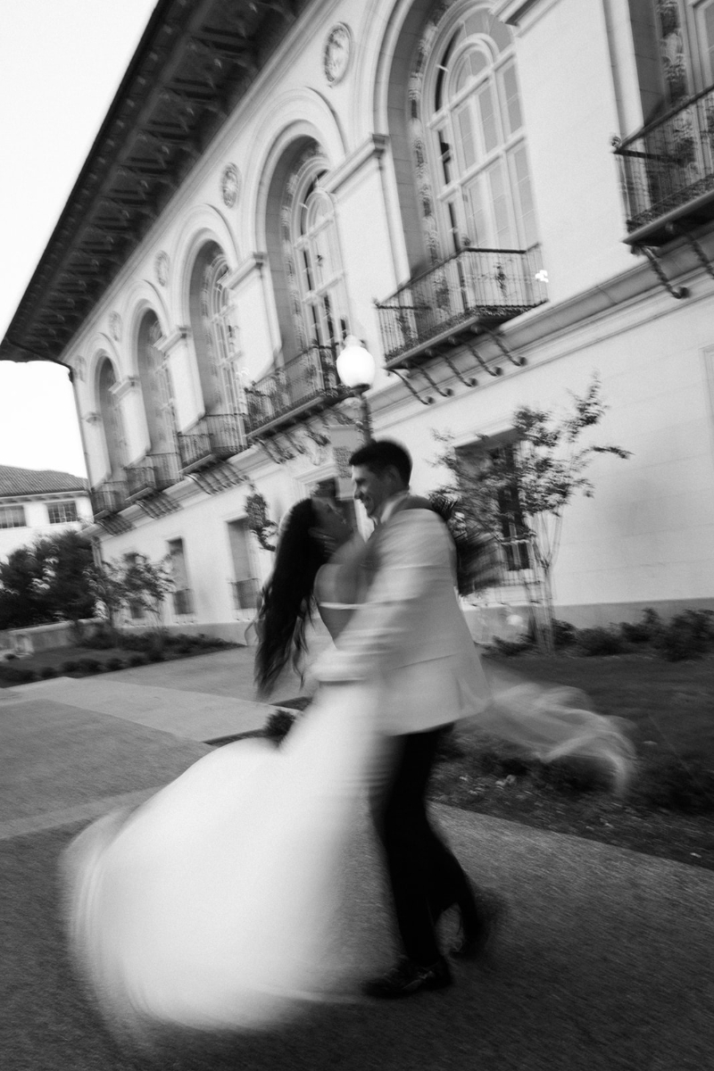 A bride and groom dance outside a building with arched windows, captured in motion blur in a black-and-white photograph.