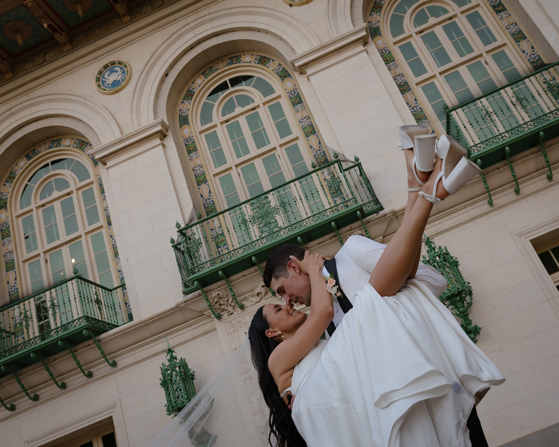 A groom lifts and holds a smiling bride in a white dress outside a historic building with arched windows and green balcony railings.