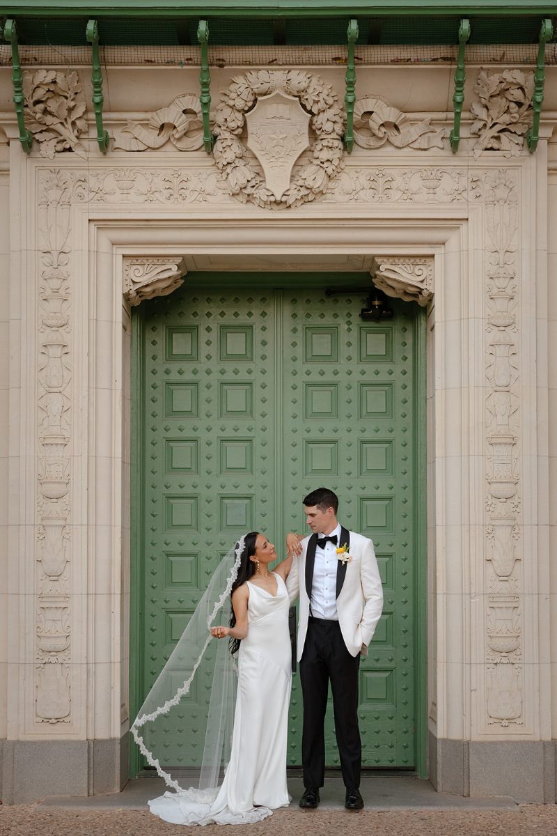 A bride and groom stand in front of a large green ornate door, dressed in formal wedding attire, looking at each during their Santa Barbara wedding.