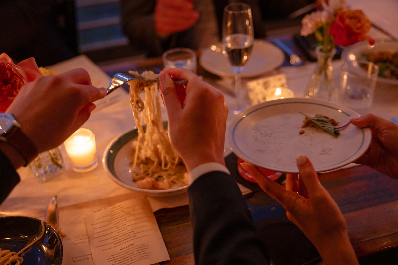 People serve pasta onto a plate at a candlelit dinner table set with wine glasses, flowers, and a menu.
