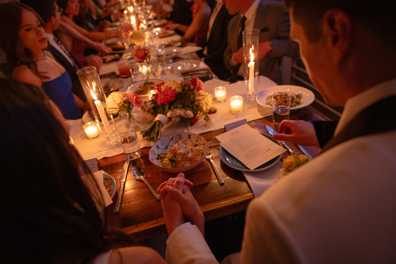 A couple holds hands at a long, candlelit dinner table with flowers, plates of food, and menus, surrounded by other formally dressed guests.