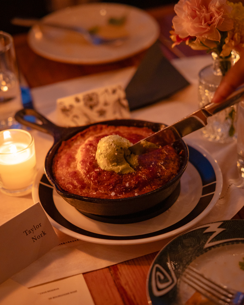 A hand places a scoop of green ice cream onto a baked dessert in a cast iron skillet on a restaurant table set with flowers, candles, and a place card reading "Taylor Nork.
