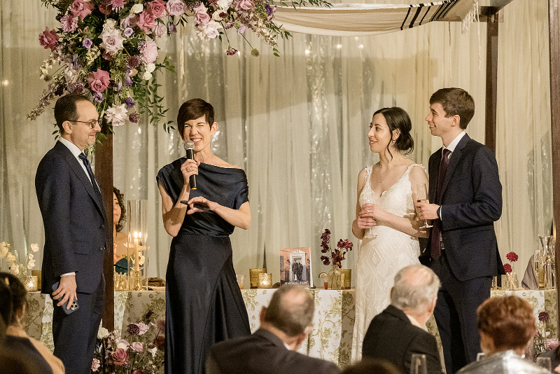 Four people stand under a flower arch at a formal event; one woman speaks into a microphone while the others listen and smile.