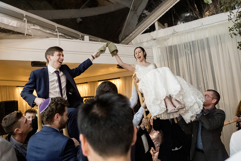 A bride and groom are lifted on chairs by guests during a wedding they planned with a destination wedding planner, each holding a cloth napkin and smiling.