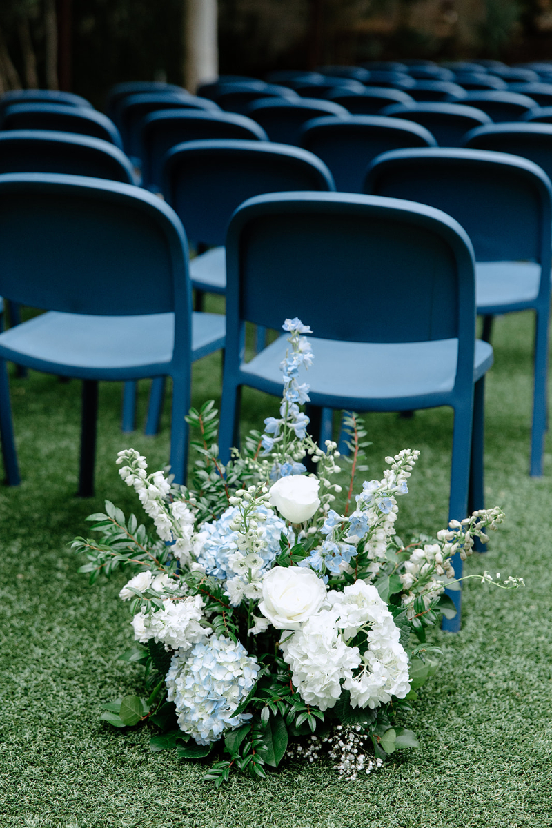 Rows of blue chairs on grass with a large arrangement of white and light blue flowers in the foreground.