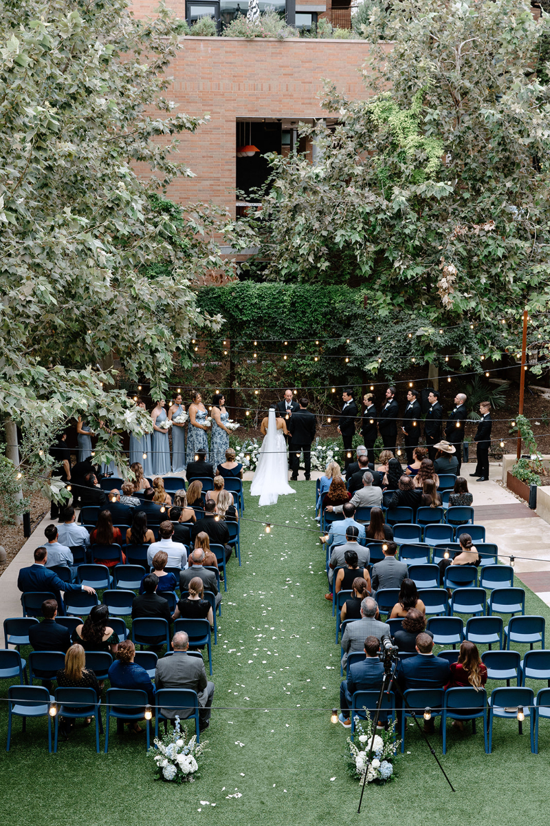 An outdoor wedding ceremony with guests seated on blue chairs, a couple standing at the altar, bridal party on each side, surrounded by trees and greenery.