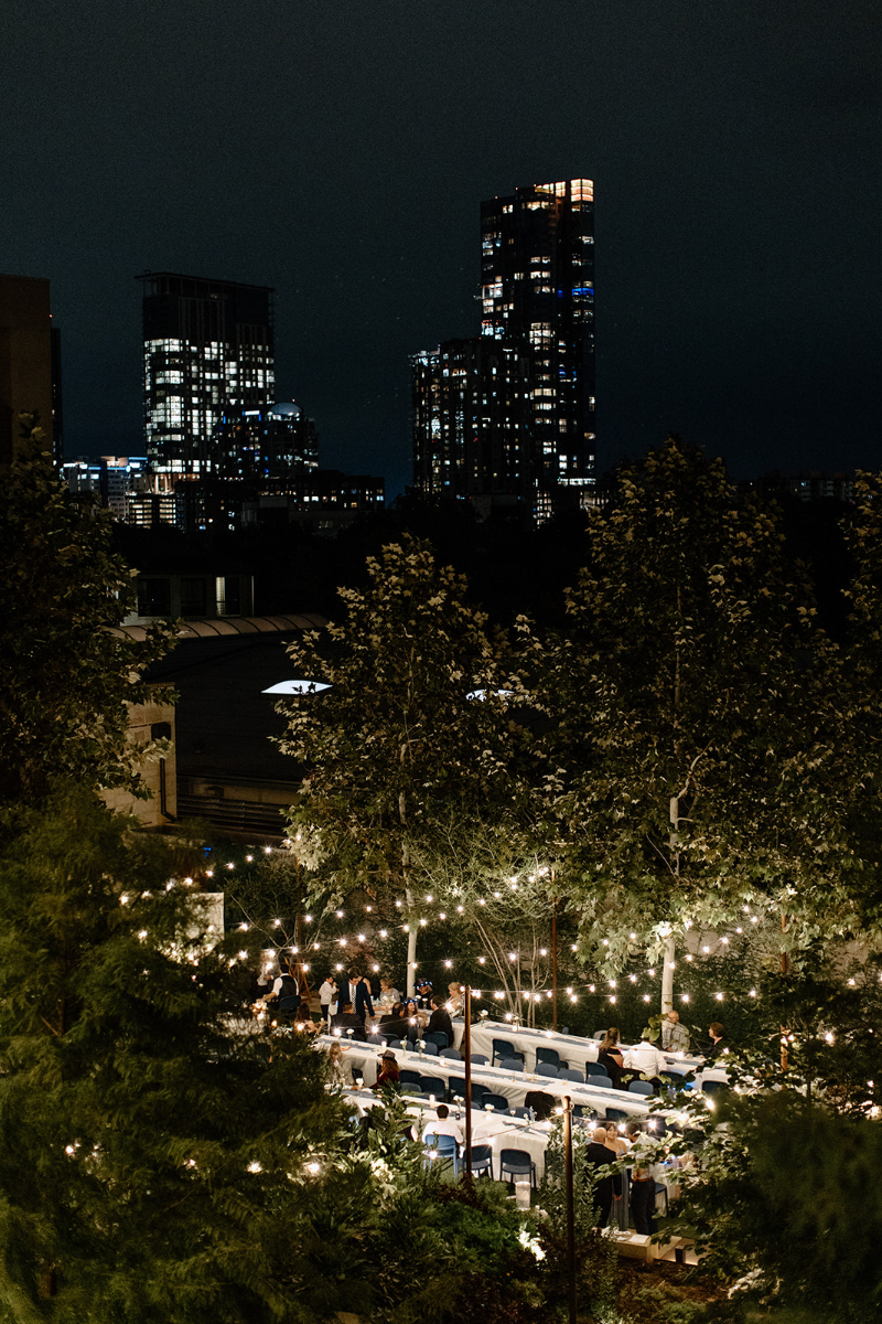 Outdoor wedding reception at Hotel Magdelena with people seated at long tables under string lights, surrounded by trees, with city skyscrapers illuminated in the background.