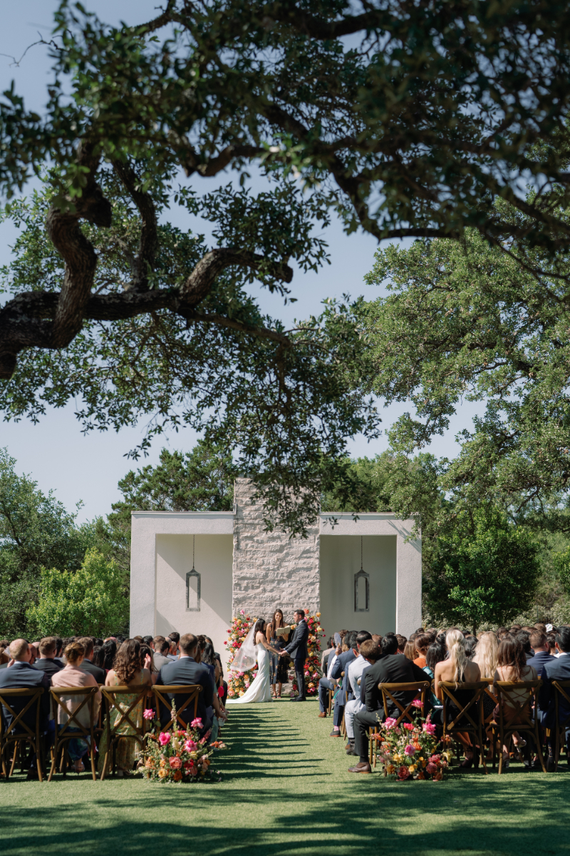A wedding ceremony takes place outdoors with a couple and officiant standing at an altar, surrounded by seated guests and greenery.